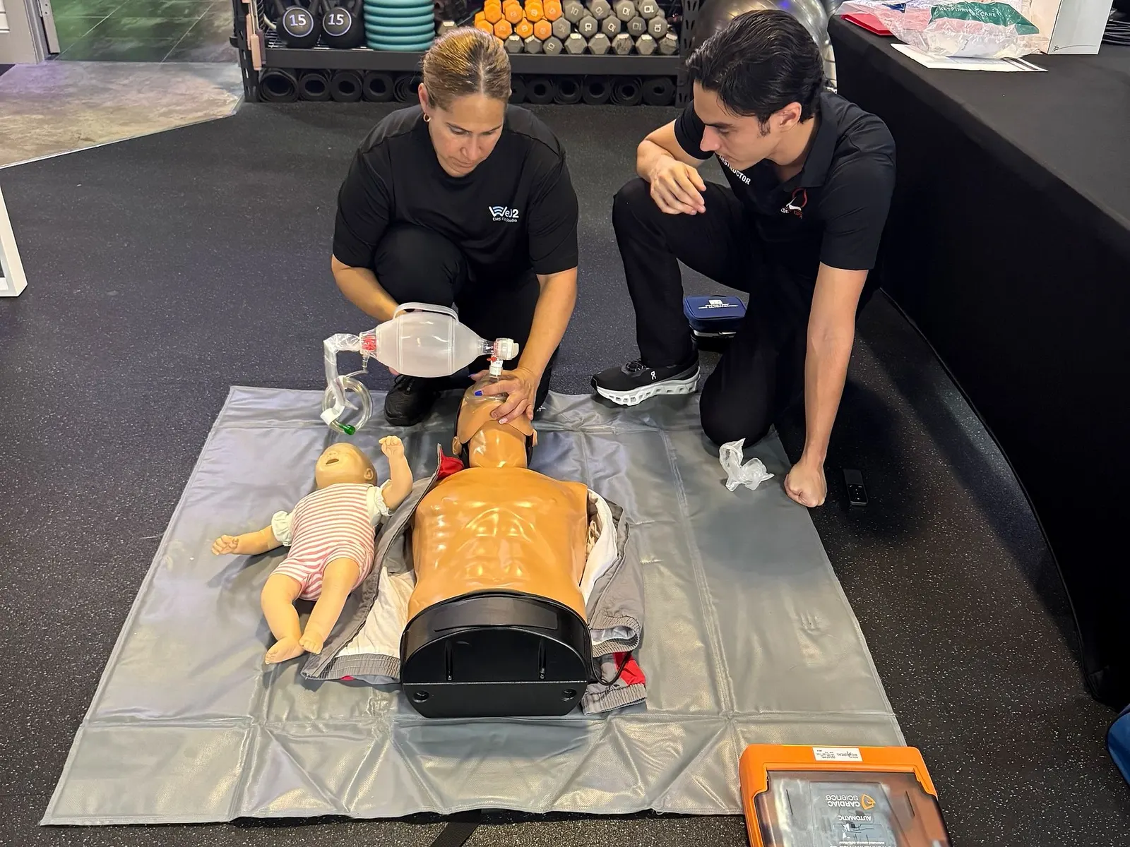 Pulse CPR instructor Peter Asencio coaching a student using a bag-mask device on an adult manikin, with an infant manikin and AED nearby